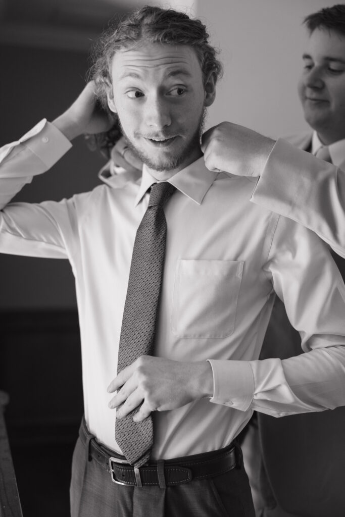 Groom adjusting tie with help from groomsmen, candid getting ready moment with natural light.