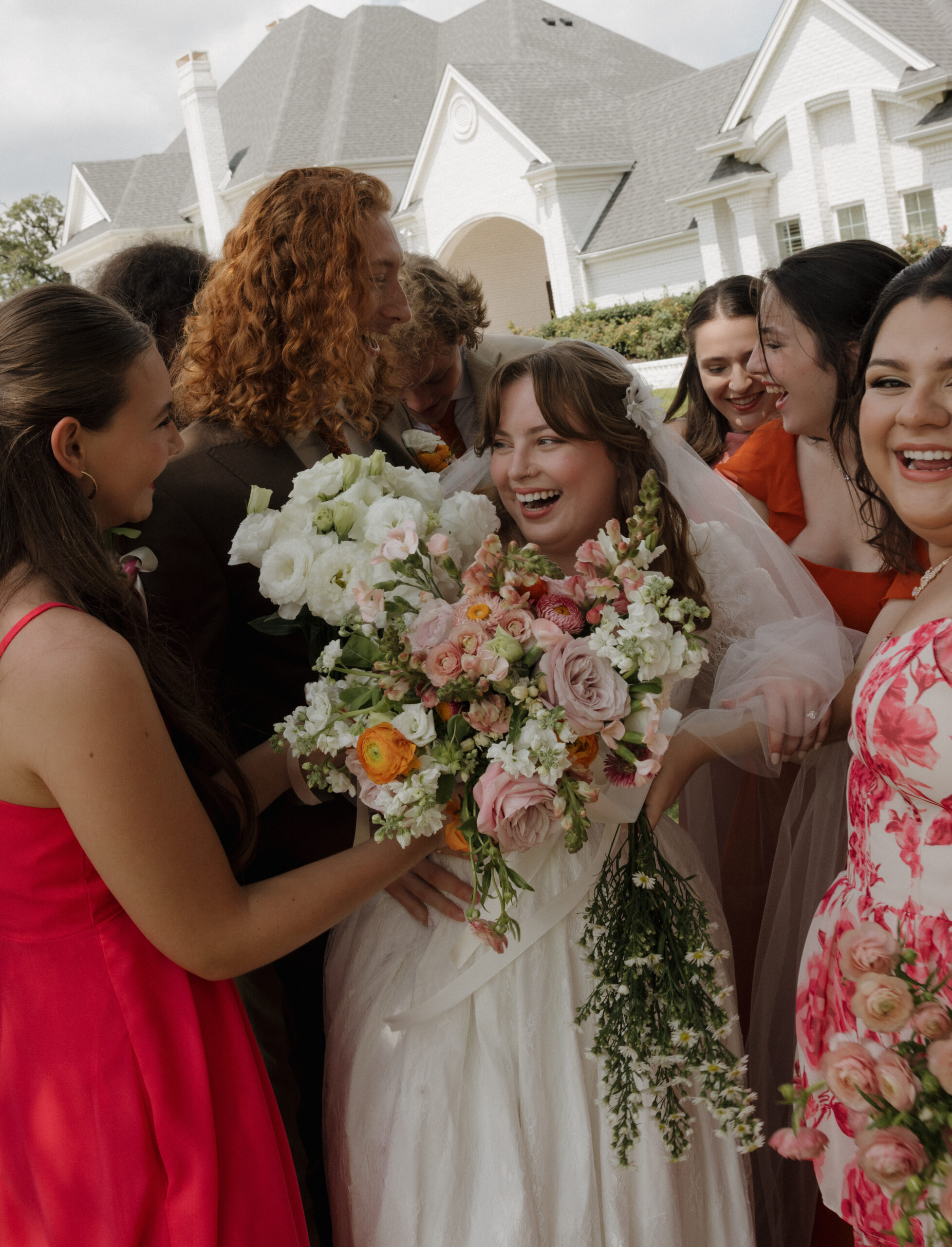 Candid moment of friends embracing newlyweds surrounded by colorful florals and sunlight.