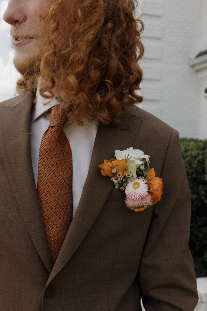 Groom wearing brown suit with orange tie and bright boutonniere of handmade florals.